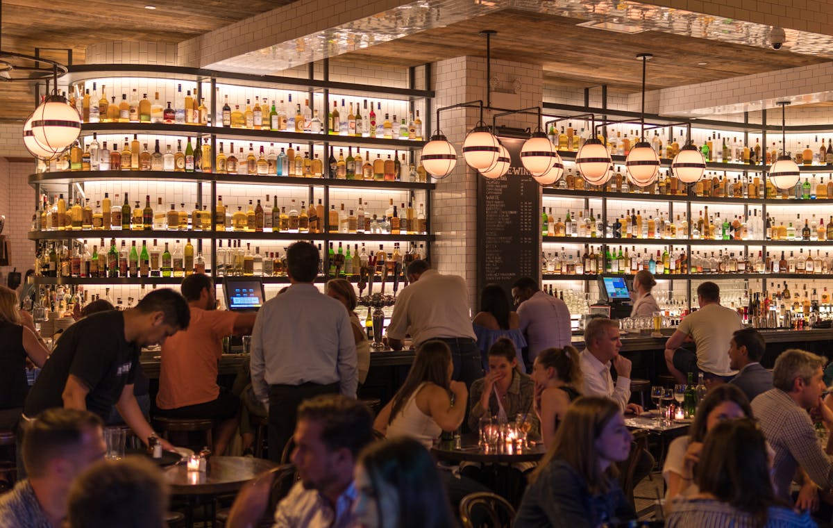 Busy restaurant bar with a wall of backlit bottles and guests seated at tables